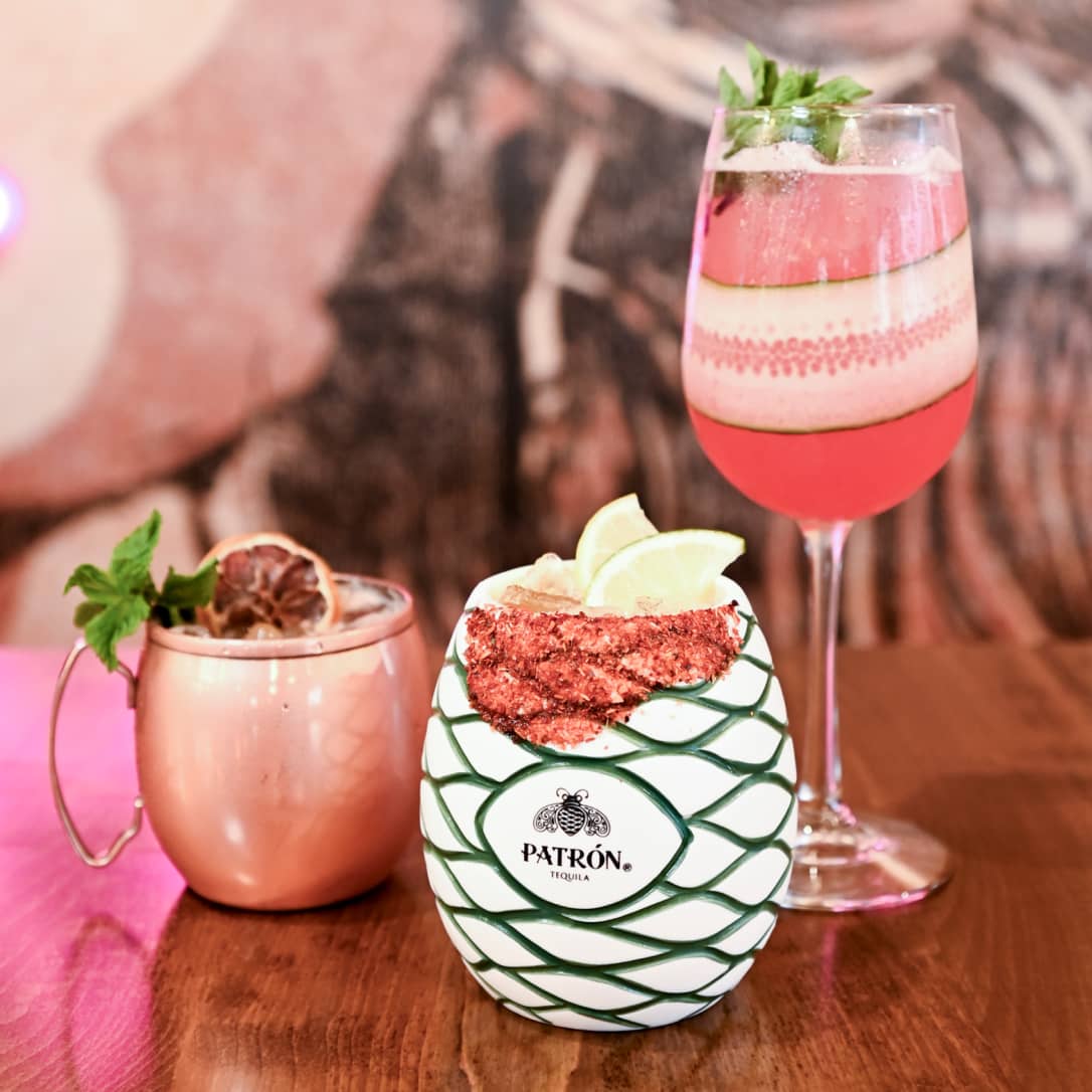Three colorful cocktails arranged on a wooden surface: a copper mug with pink drink and mint garnish on the left, a pineapple-shaped white ceramic mug with green geometric pattern and Patrón tequila logo containing a red-rimmed drink with lime wedges in the center, and a tall glass with layered pink and clear liquid garnished with mint on the right.