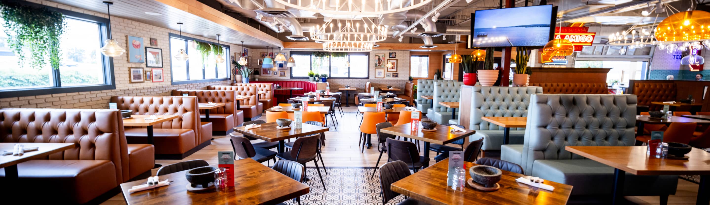 Interior view of a modern casual restaurant with mixed seating including brown leather booth banquettes along brick walls and gray upholstered booth seating. The space features wooden tables with black chairs, exposed ceiling beams with hanging pendant lights, large windows providing natural light, and decorative elements including hanging plants and framed artwork on brick walls. Television screens are mounted throughout the dining area, and the space has an industrial-modern aesthetic with warm lighting.