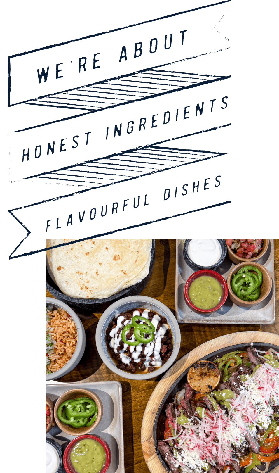 Overhead view of Mexican food spread on a wooden table with a decorative banner reading "WE'RE ABOUT HONEST INGREDIENTS FLAVOURFUL DISHES." The meal includes flour tortillas in a cast iron pan, rice and beans in bowls, a dish with sour cream and jalapeños, various salsas and guacamole in small bowls, and a large wooden bowl containing what appears to be fajitas with peppers, onions, and cheese.